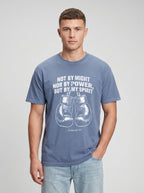 A man wearing a slate blue graphic t-shirt with a white boxing design and the quote 'NOT BY MIGHT NOR BY POWER, BUT BY MY SPIRIT' paired with light wash jeans, against a neutral gray background.