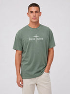 A man wearing a sage green t-shirt with a white cross and the gothic-style text 'jesus saves' across the chest, paired with light beige pants, against a light gray studio background.