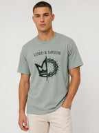 A man wearing a light sage green t-shirt with a black graphic of a crown of thorns and the text 'LORD & SAVIOR' printed on the chest, paired with off-white pants, against a plain light gray background.