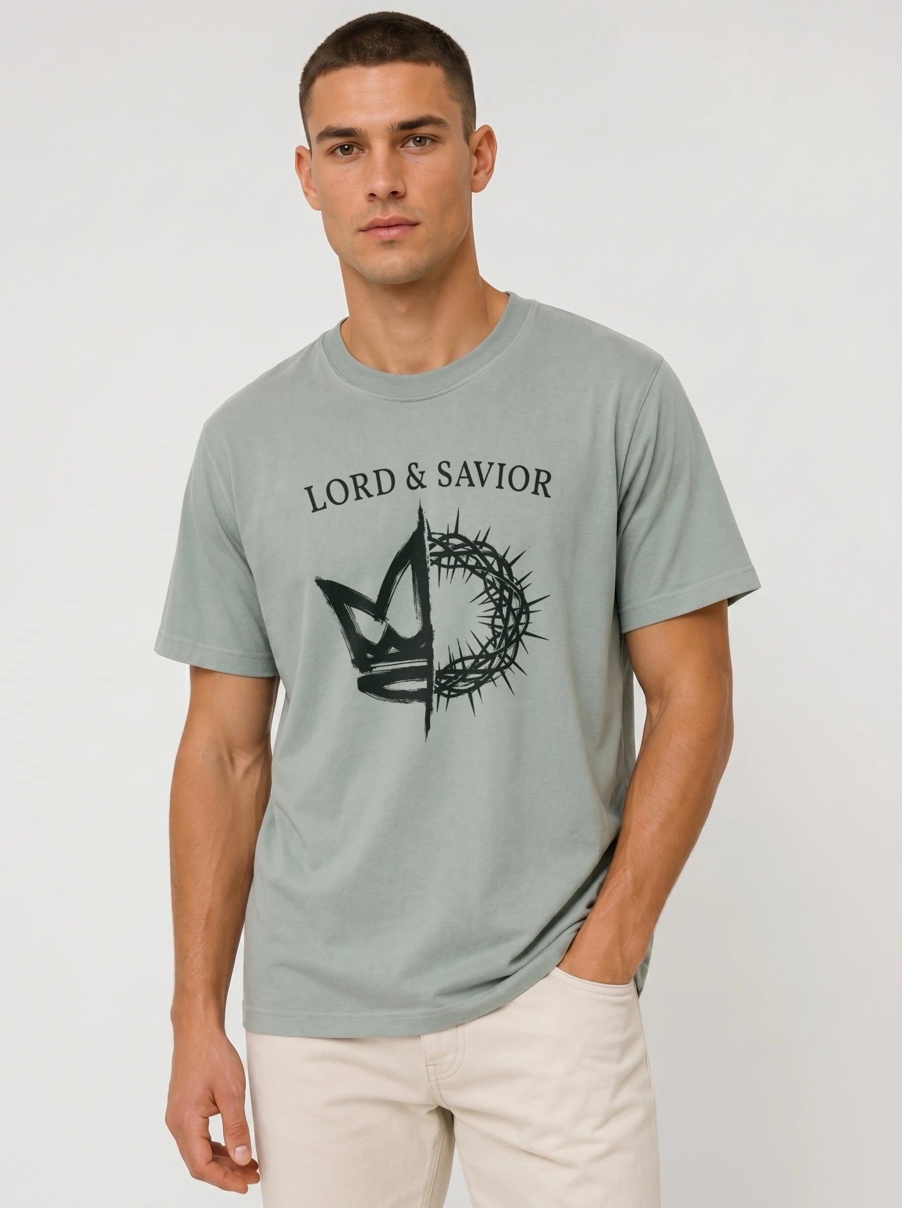 A man wearing a light sage green t-shirt with a black graphic of a crown of thorns and the text 'LORD & SAVIOR' printed on the chest, paired with off-white pants, against a plain light gray background.
