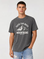 A man wearing a dark gray graphic t-shirt with a white mountain design and the text 'FAITH CAN MOVE MOUNTAINS' and 'Matthew 17:20', paired with light blue jeans, against a light gray studio background.