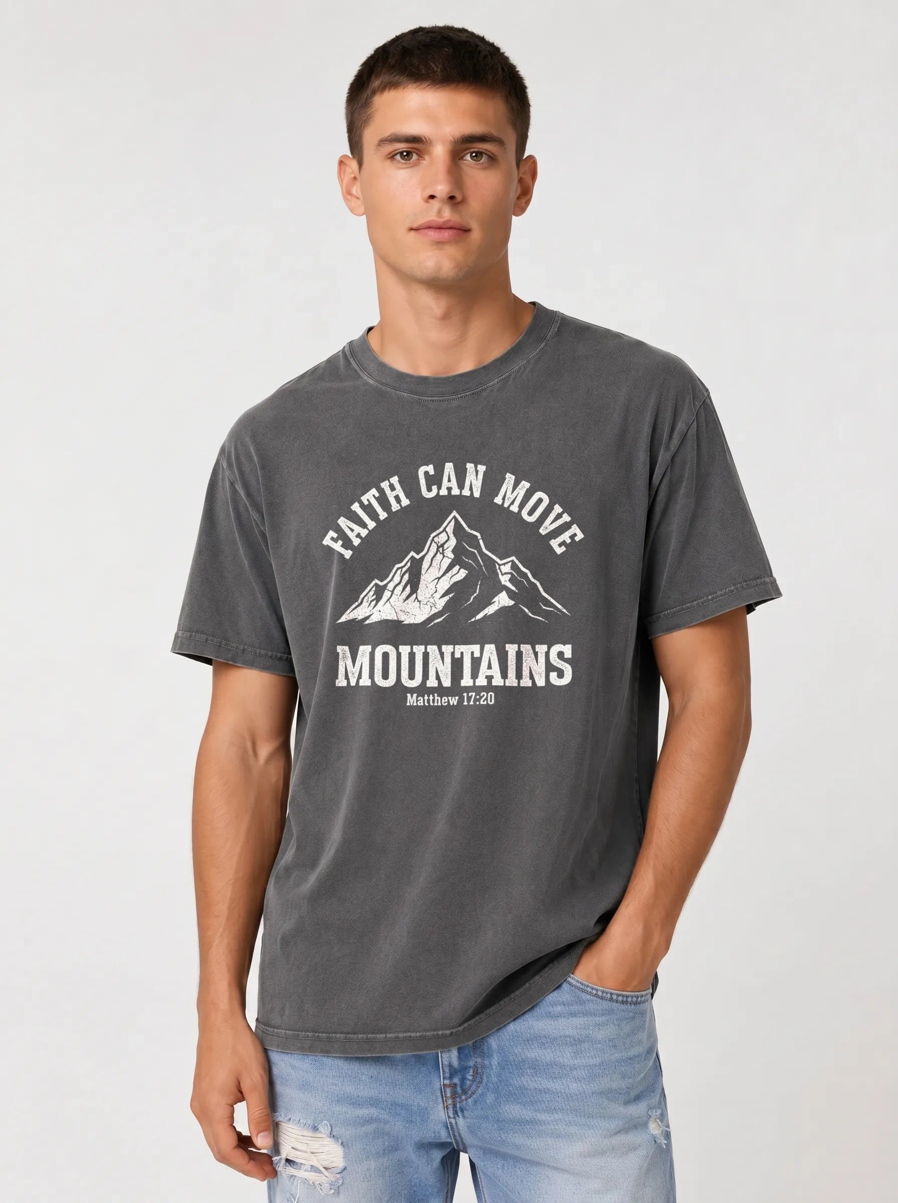 A man wearing a dark gray graphic t-shirt with a white mountain design and the text 'FAITH CAN MOVE MOUNTAINS' and 'Matthew 17:20', paired with light blue jeans, against a light gray studio background.