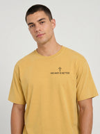 A man wearing a mustard yellow t-shirt with the text 'HIS WAY IS BETTER' and a small cross graphic printed on the chest, photographed against a light gray studio background.