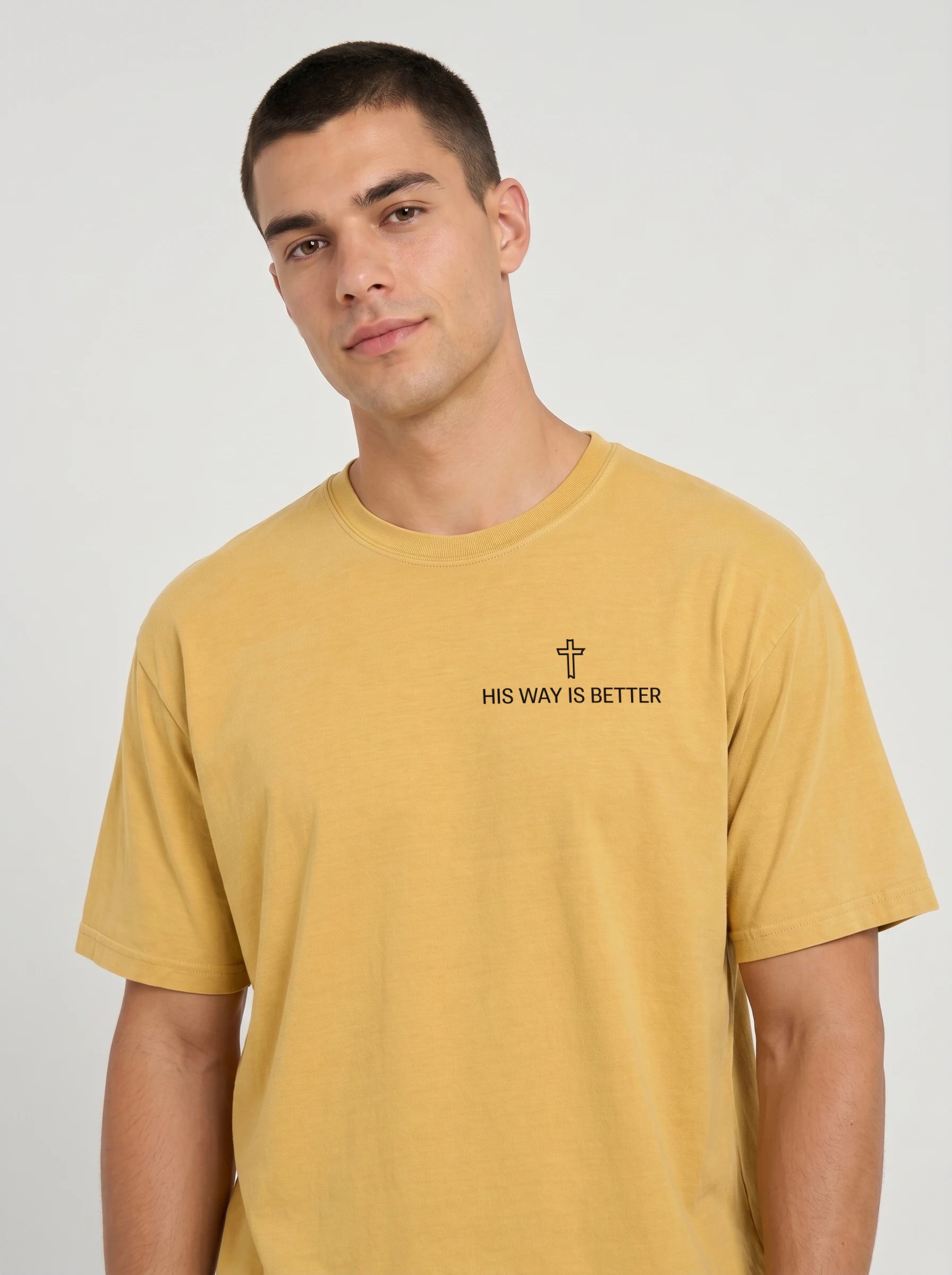 A man wearing a mustard yellow t-shirt with the text 'HIS WAY IS BETTER' and a small cross graphic printed on the chest, photographed against a light gray studio background.
