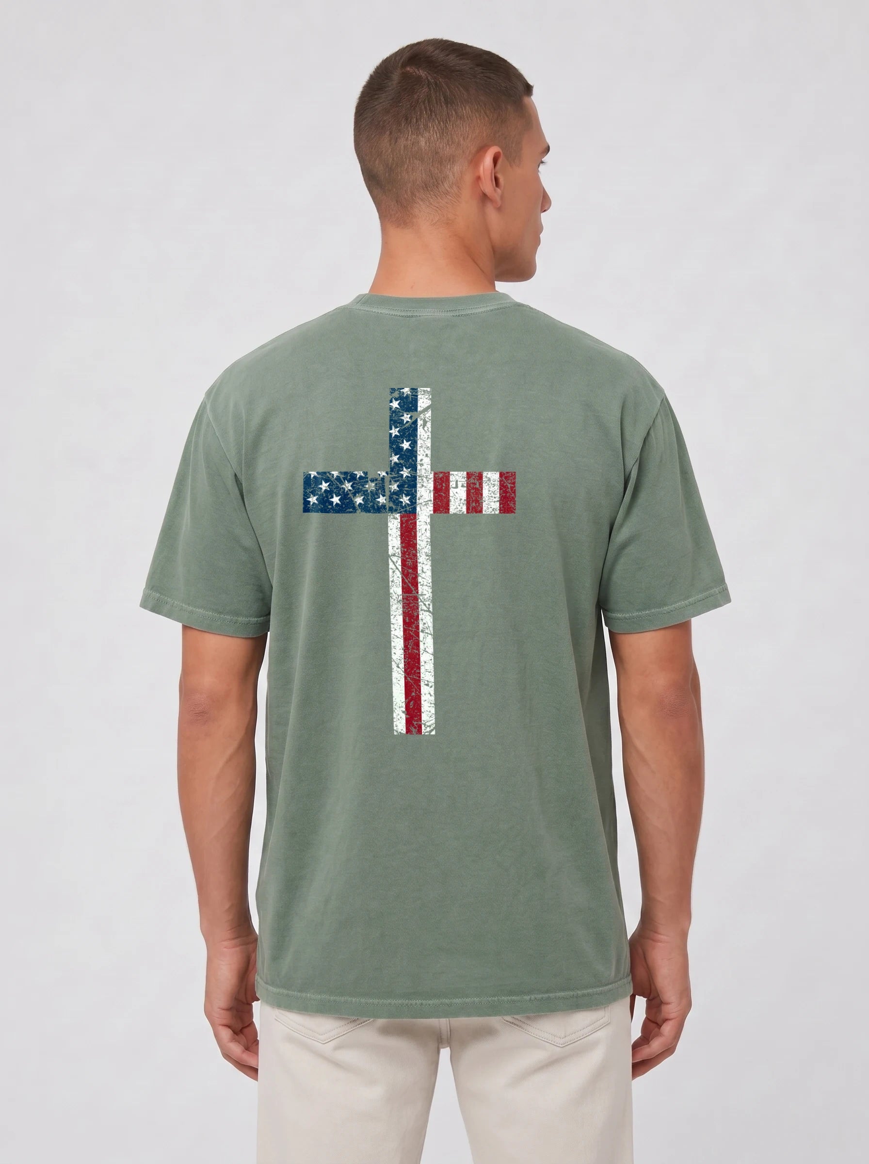 A man viewed from the back wearing a faded sage green t-shirt with a large distressed cross graphic in the American flag colors (blue, red, and white) on the back, paired with light beige pants, against a light gray studio background.