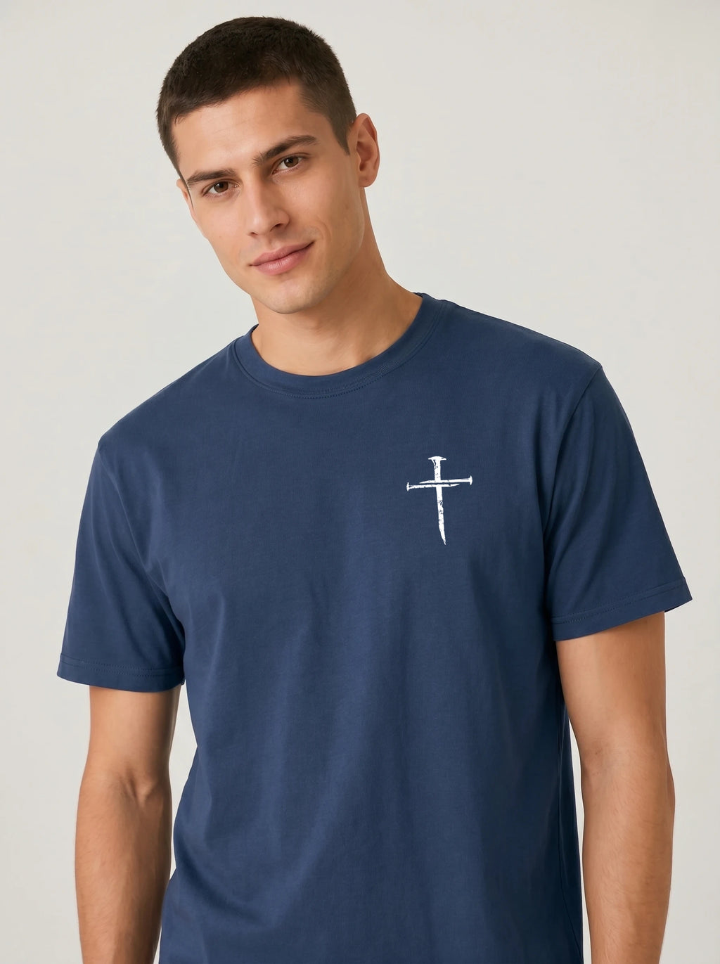 A man wearing a navy blue crewneck t-shirt with a small white nail cross graphic printed on the left chest, photographed against a light gray studio background.