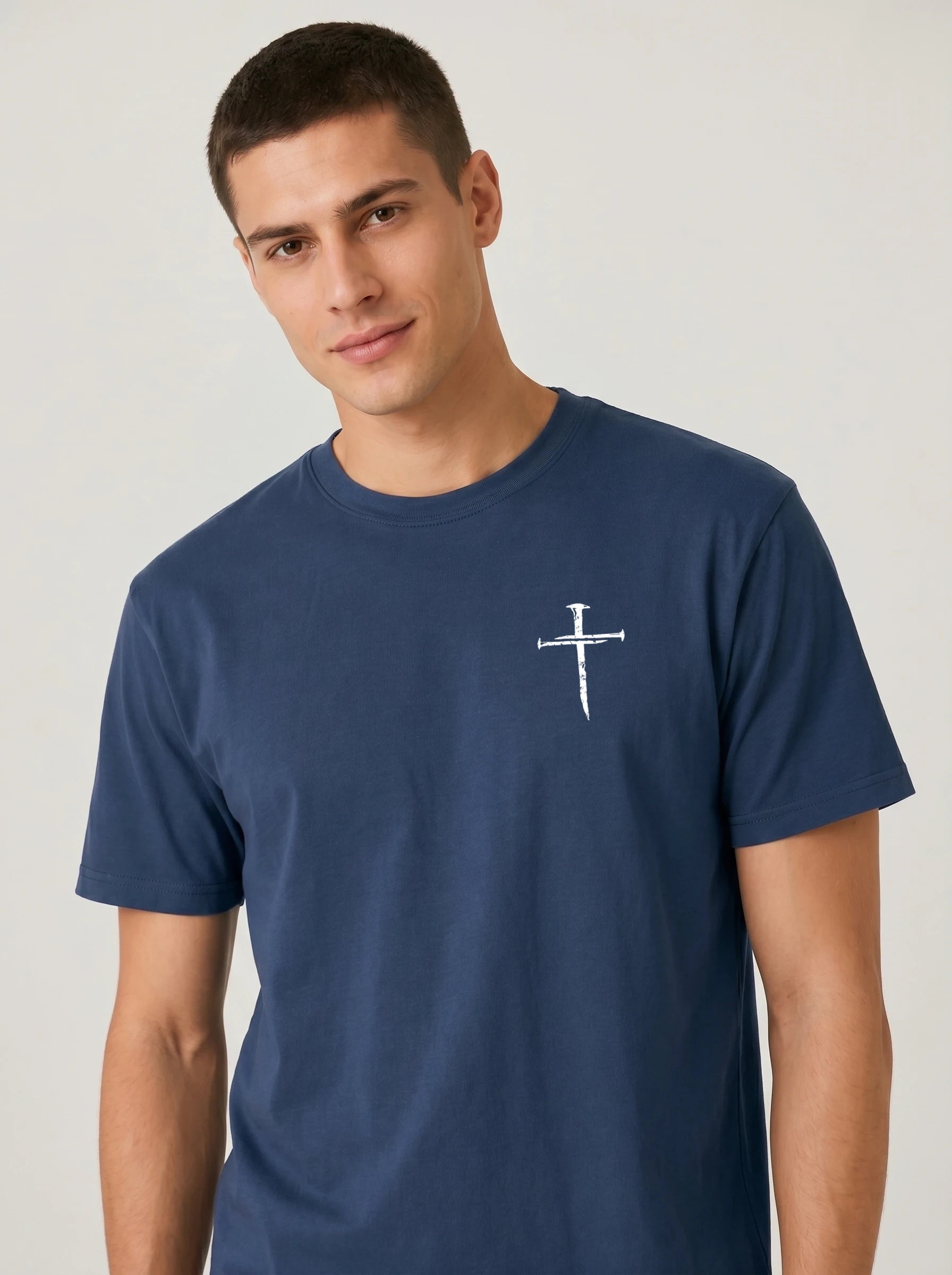 A man wearing a navy blue crewneck t-shirt with a small white nail cross graphic printed on the left chest, photographed against a light gray studio background.