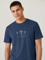 A man wearing a navy blue crewneck t-shirt with a white graphic of three crosses and the text 'FAITH OVER FEAR' printed on the chest, set against a plain light gray background.