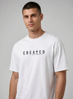 A man wearing a white crewneck t-shirt with the text 'CREATED WITH A PURPOSE' printed in black across the chest, photographed against a neutral gray studio background.