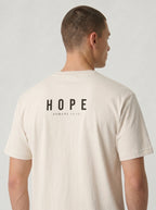A man viewed from the back wearing a cream-colored t-shirt with 'HOPE ROMANS 15:13' printed in black text on the back, set against a plain light gray background.