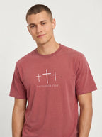 A man wearing a dusty rose short-sleeve t-shirt with a white graphic of three crosses and the text 'FAITH OVER FEAR' printed on the chest, set against a plain light gray background.