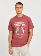 A man wearing a dusty rose short-sleeve t-shirt with a white graphic print of boxing gloves and the biblical quote 'NOT BY MIGHT NOR BY POWER, BUT BY MY SPIRIT', paired with light beige trousers, against a plain light gray background.