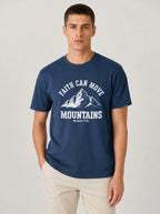 A man wearing a navy blue crewneck t-shirt with a white graphic print that reads 'FAITH CAN MOVE MOUNTAINS' and 'Mountain 17:20' below it, paired with beige pants, against a light gray studio background.