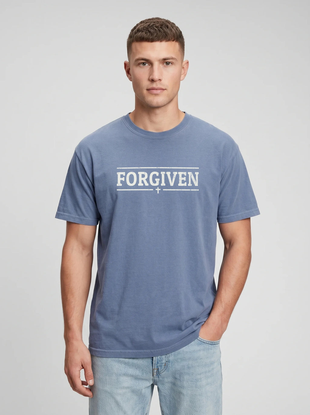 A man wearing a slate blue t-shirt with 'FORGIVEN' printed in white across the chest, paired with light-wash jeans, against a neutral gray studio background.