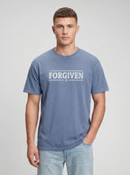 A man wearing a slate blue t-shirt with 'FORGIVEN' printed in white across the chest, paired with light-wash jeans, against a neutral gray studio background.
