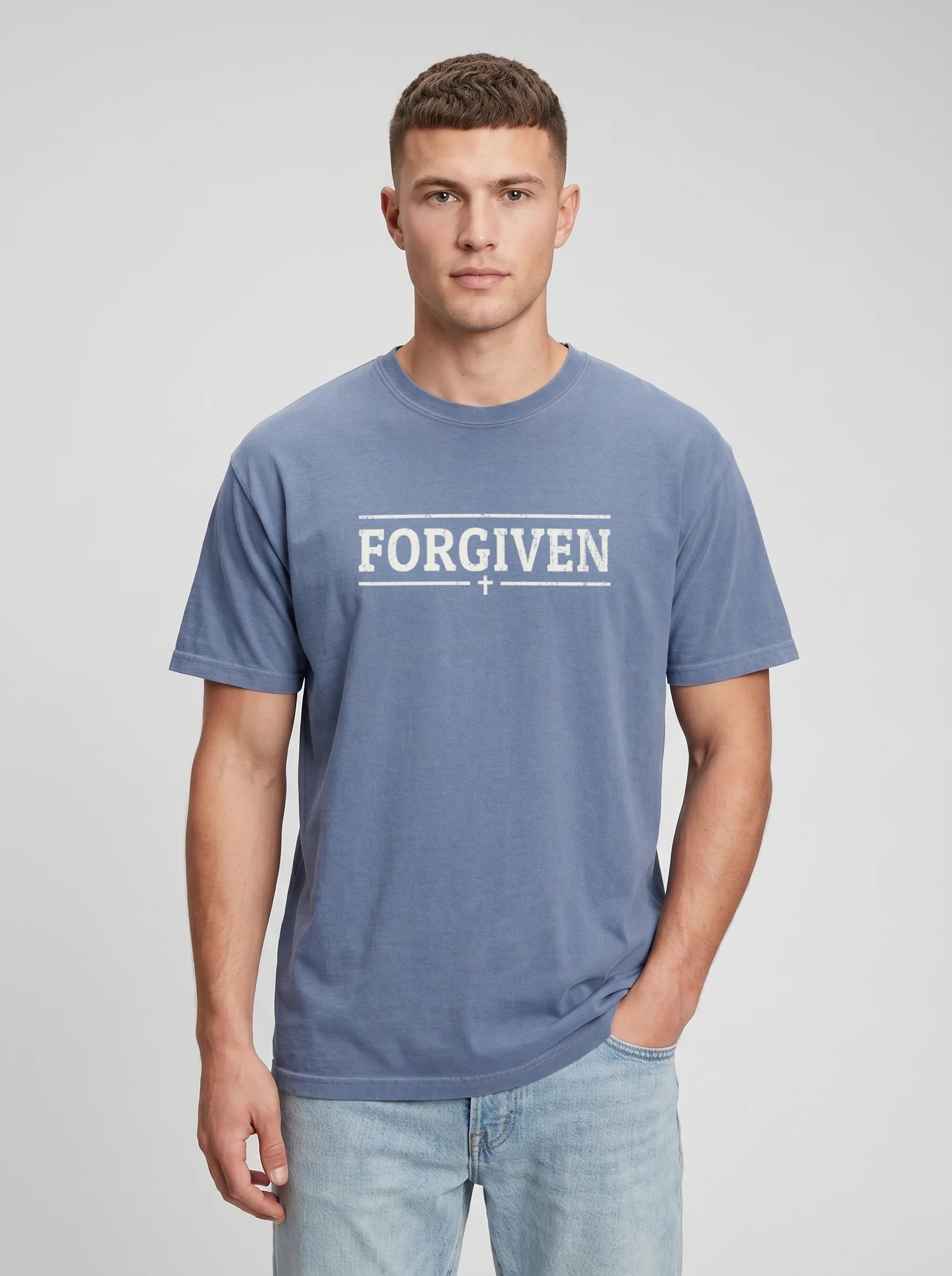 A man wearing a slate blue t-shirt with 'FORGIVEN' printed in white across the chest, paired with light-wash jeans, against a neutral gray studio background.