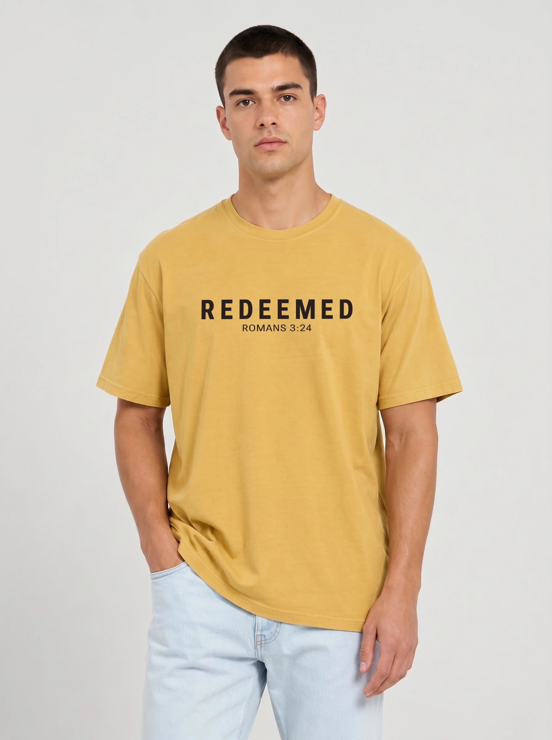 A man wearing a mustard yellow t-shirt with the text 'REDEEMED ROMANS 3:24' printed in black across the chest, paired with light blue jeans, against a plain light gray background.
