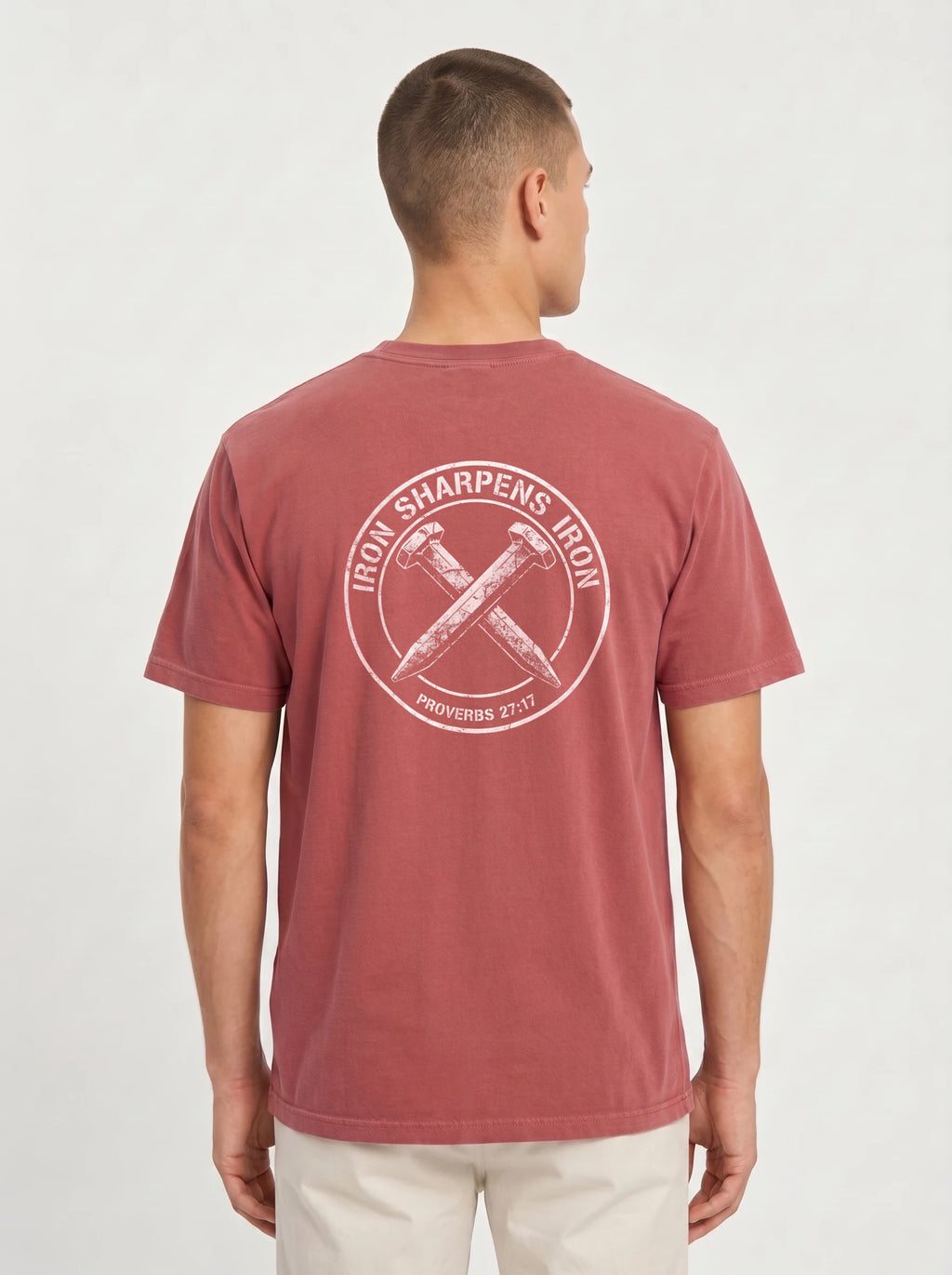 A man viewed from the back wearing a muted red crewneck t-shirt with a large white circular graphic on the back that reads 'IRON SHARPENS IRON' and 'PROVERBES 27:17' around crossed hammers. He is also wearing light beige pants against a clean, light gray studio background.
