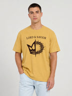 A man wearing a mustard yellow t-shirt with a black graphic of a crown of thorns and the text 'LORD & SAVIOR' printed on the chest, paired with light blue jeans, against a plain light gray background.