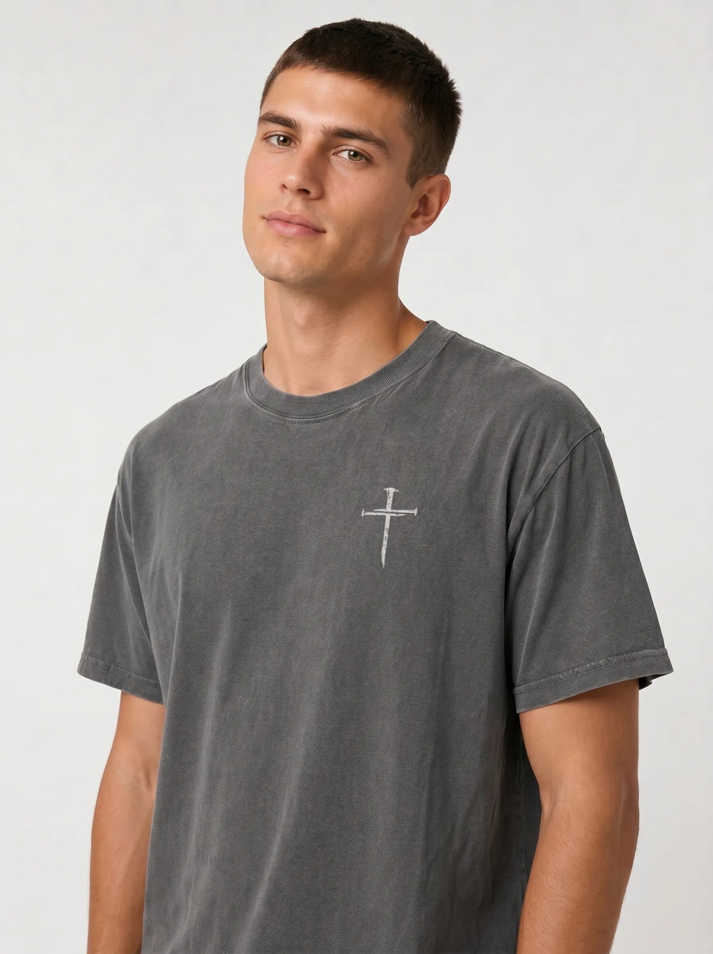 A man wearing a dark gray washed t-shirt with a small cross printed on the chest, photographed against a light gray studio background.