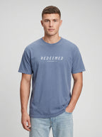 A man wearing a steel blue Comfort Colors t-shirt with "REDEEMED" in bold white capital letters and "EPHESIANS 1:7" underneath, paired with light denim jeans, against a plain light gray studio background.