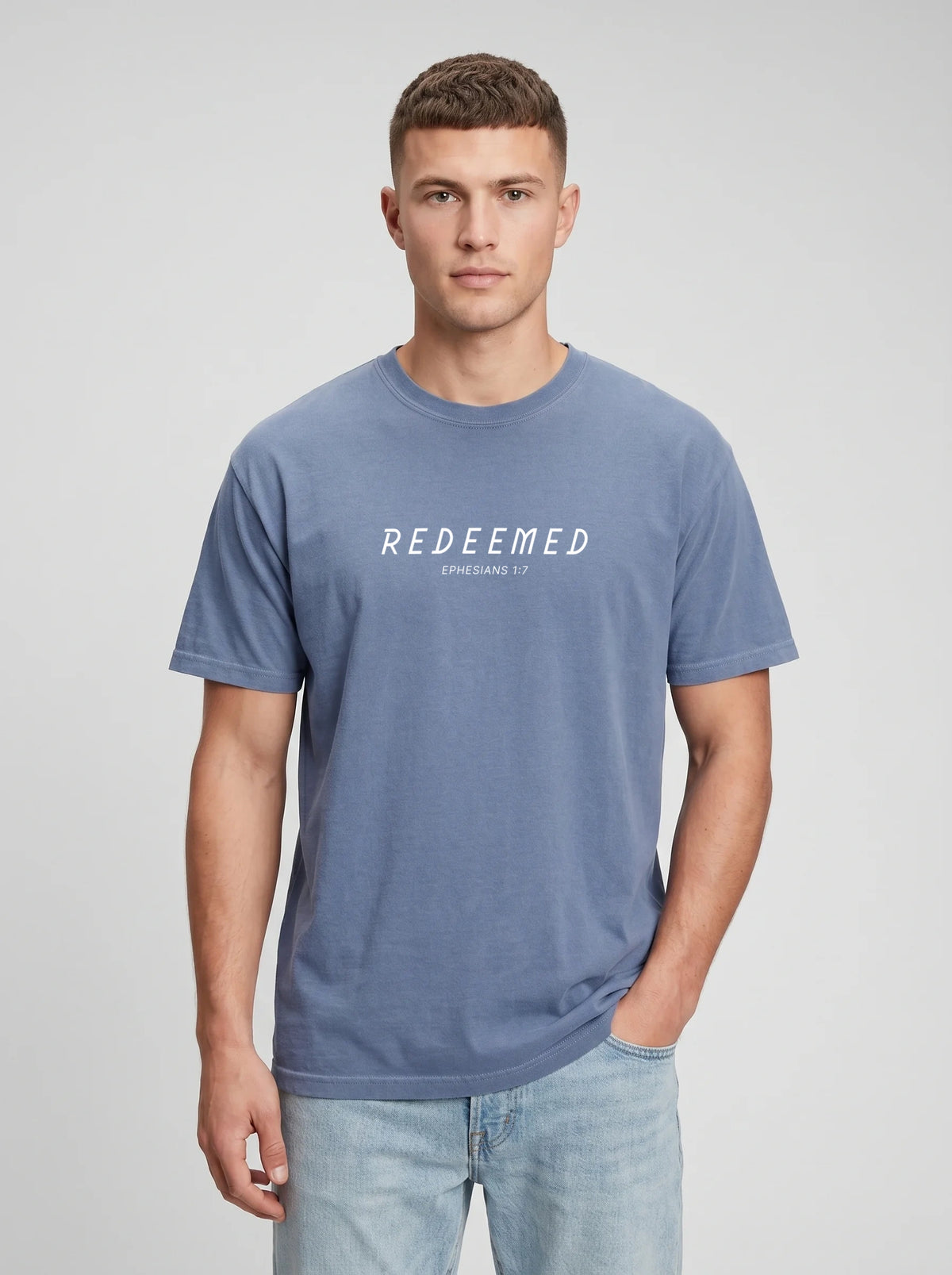A man wearing a steel blue Comfort Colors t-shirt with "REDEEMED" in bold white capital letters and "EPHESIANS 1:7" underneath, paired with light denim jeans, against a plain light gray studio background.
