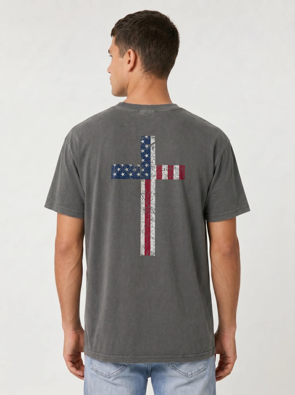 A man viewed from the back wearing a washed dark gray t-shirt with a distressed American flag cross graphic on the back, paired with light blue jeans, against a light gray studio background.