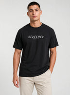 A man wearing a black Comfort Colors t-shirt with "REDEEMED" in bold white capital letters and "EPHESIANS 1:7" printed beneath it across the chest, paired with beige pants against a plain light gray studio background.