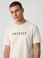 A man wearing a light beige crewneck t-shirt with the text 'CREATED WITH A PURPOSE' printed in black across the chest, set against a plain light gray background.