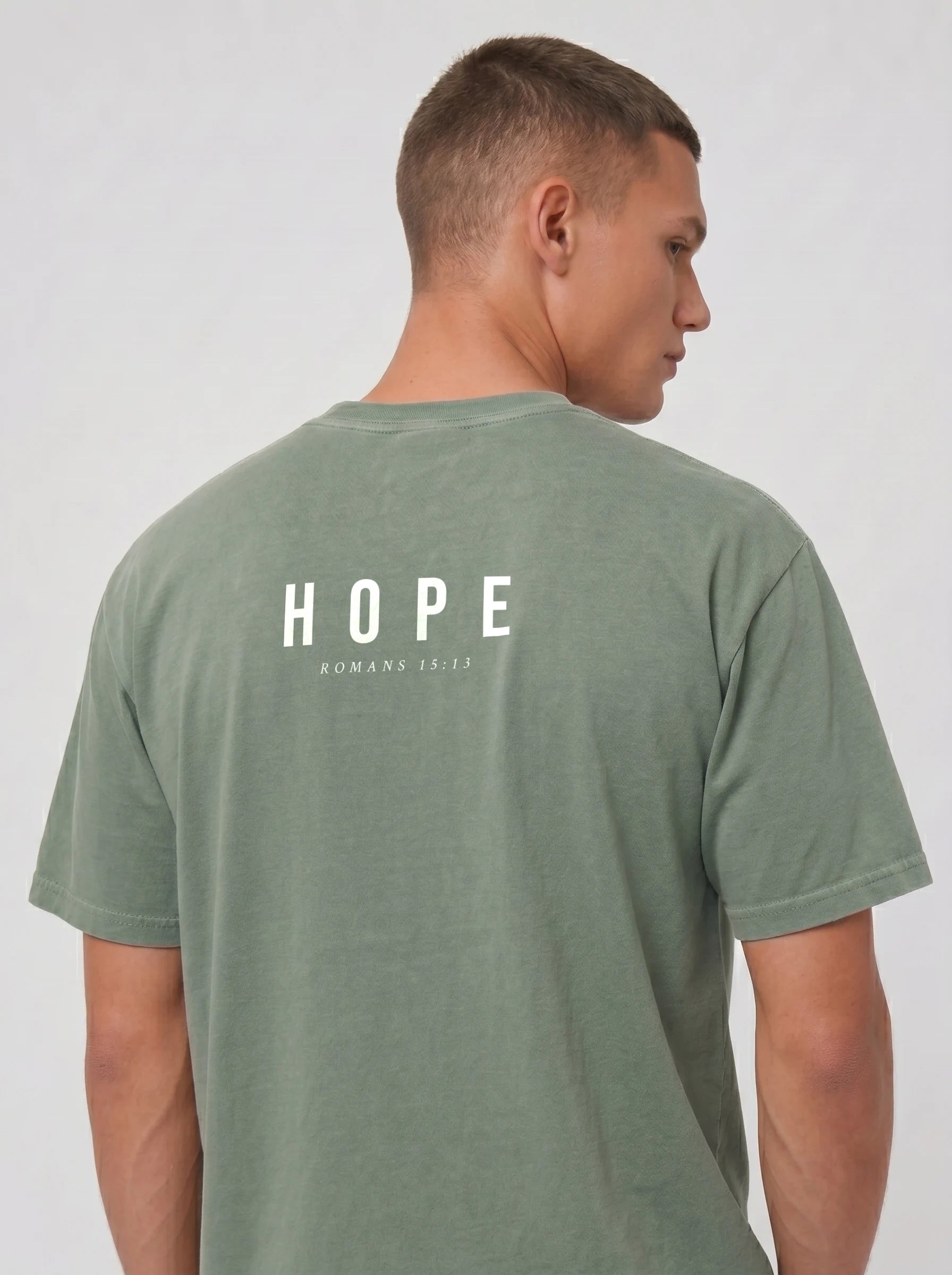 A man shown from the back wearing a sage green t-shirt with 'HOPE' and 'ROMANS 15:13' printed in white on the back, against a light gray studio background.