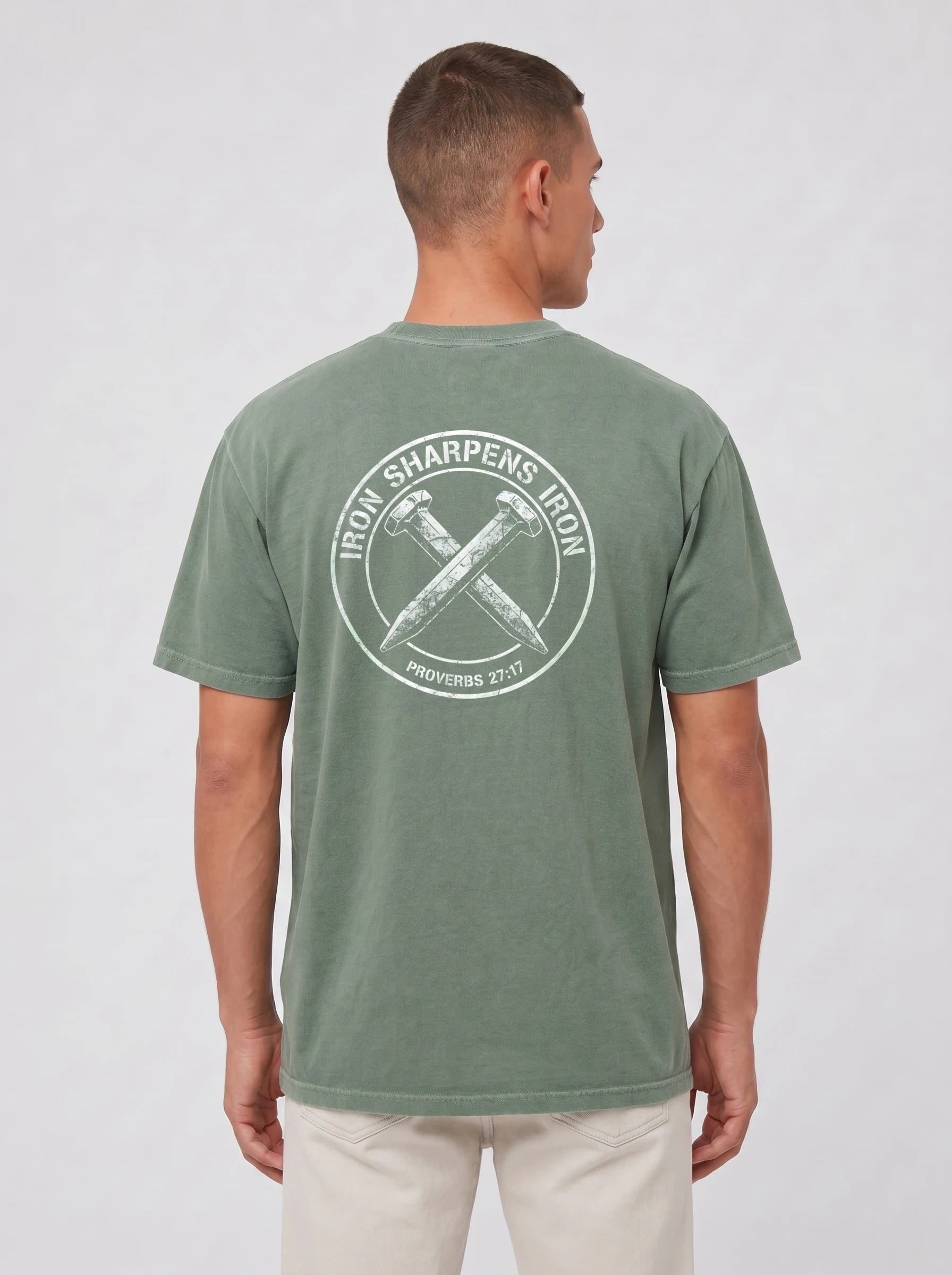 A man viewed from the back wearing a heathered sage green t-shirt with a large white circular graphic on the back with the text 'IRON SHARPENS IRON' and 'PROVERBS 27:17' surrounding an illustration of two crossed nails.