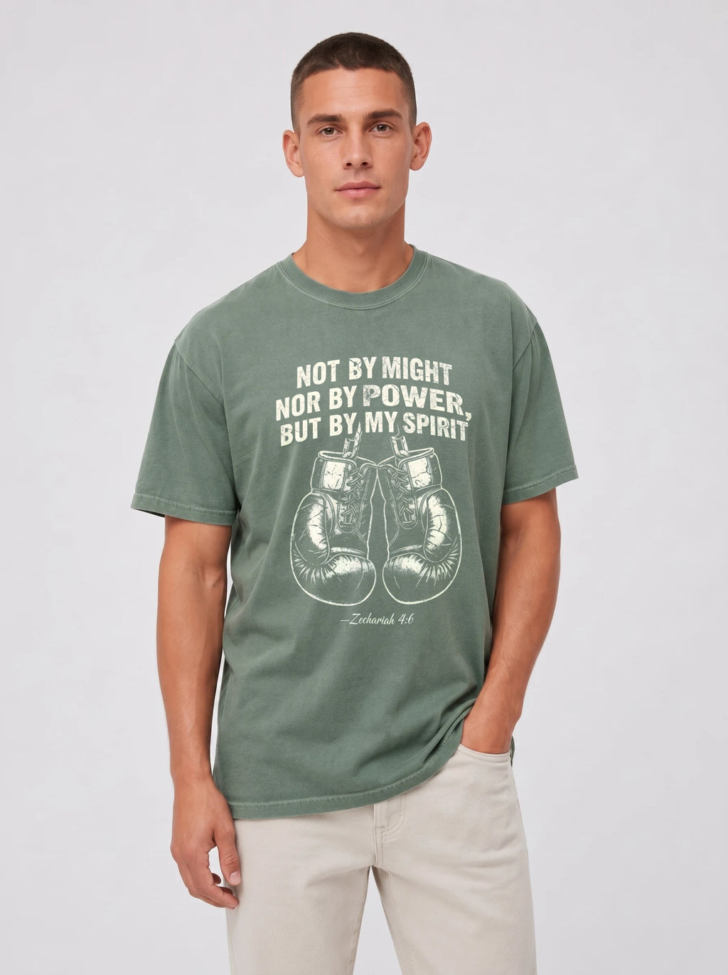 A man wearing a sage green graphic t-shirt with a white boxing theme and motivational quote, paired with beige pants, against a light gray studio background.