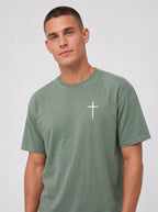 A man wearing a muted sage green crewneck t-shirt with a small white cross graphic on the left chest, photographed against a light gray studio background.