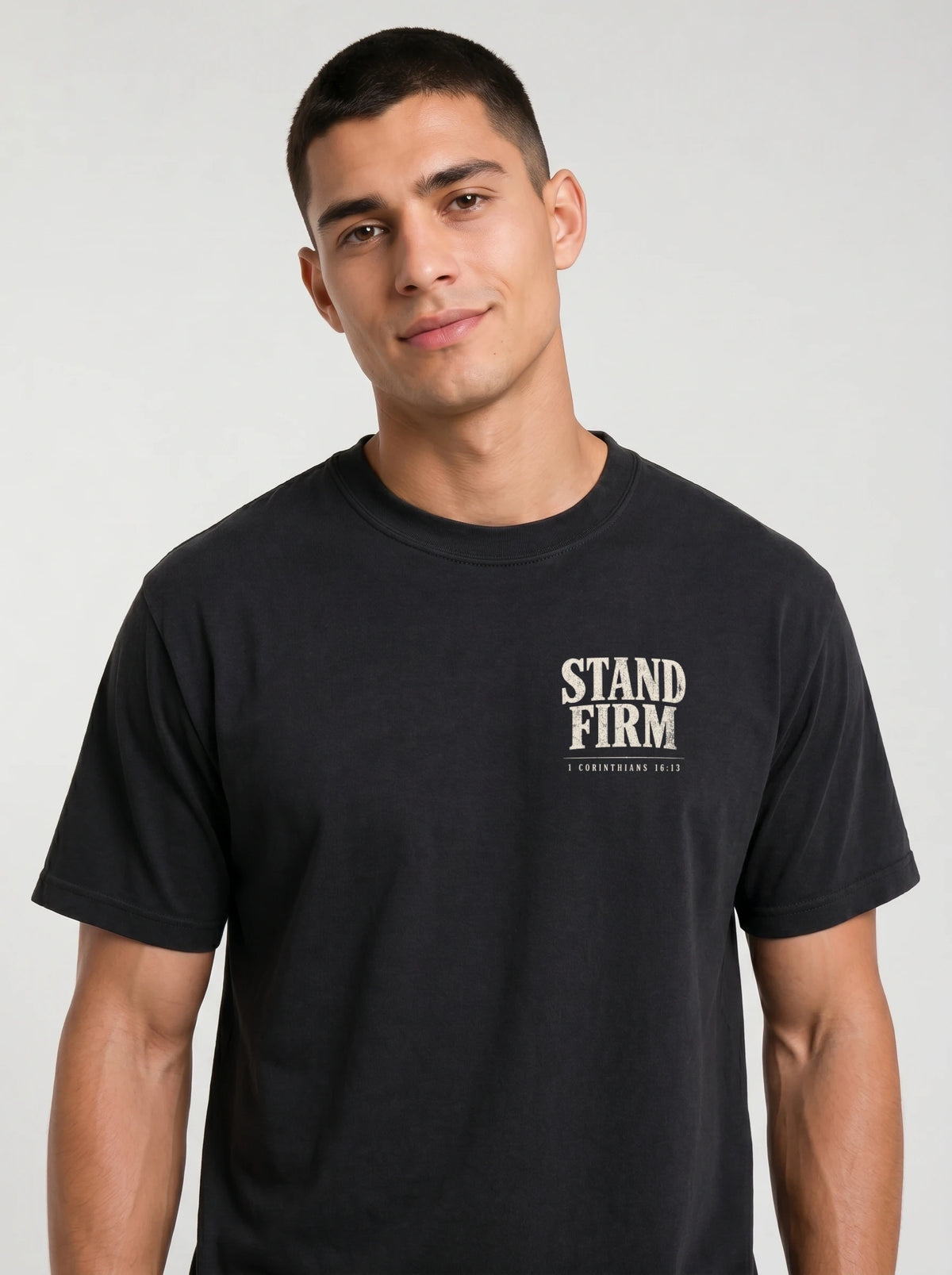 A man wearing a black short-sleeve t-shirt with a white graphic printed on the chest that say ´STAND FIRM´, photographed against a neutral light gray background.
