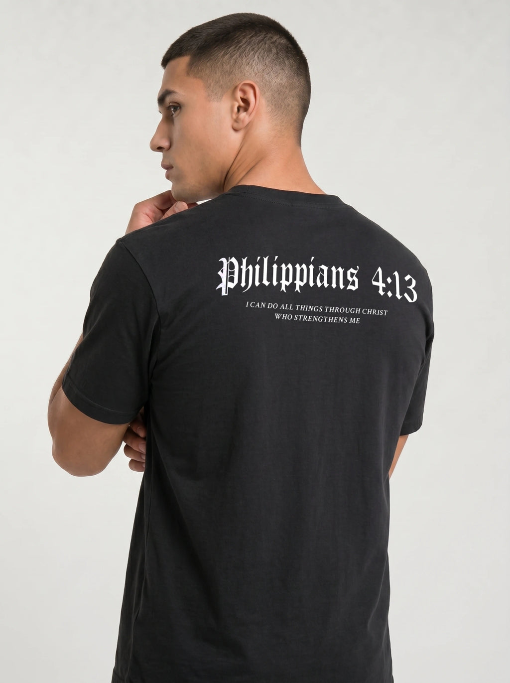 A man wearing a black t-shirt, viewed from the back. The shirt features the text 'Philippians 4:13' in a white Gothic font, with the subtitle 'I CAN DO ALL THINGS THROUGH CHRIST WHO STRENGTHENS ME' printed below in smaller white text.