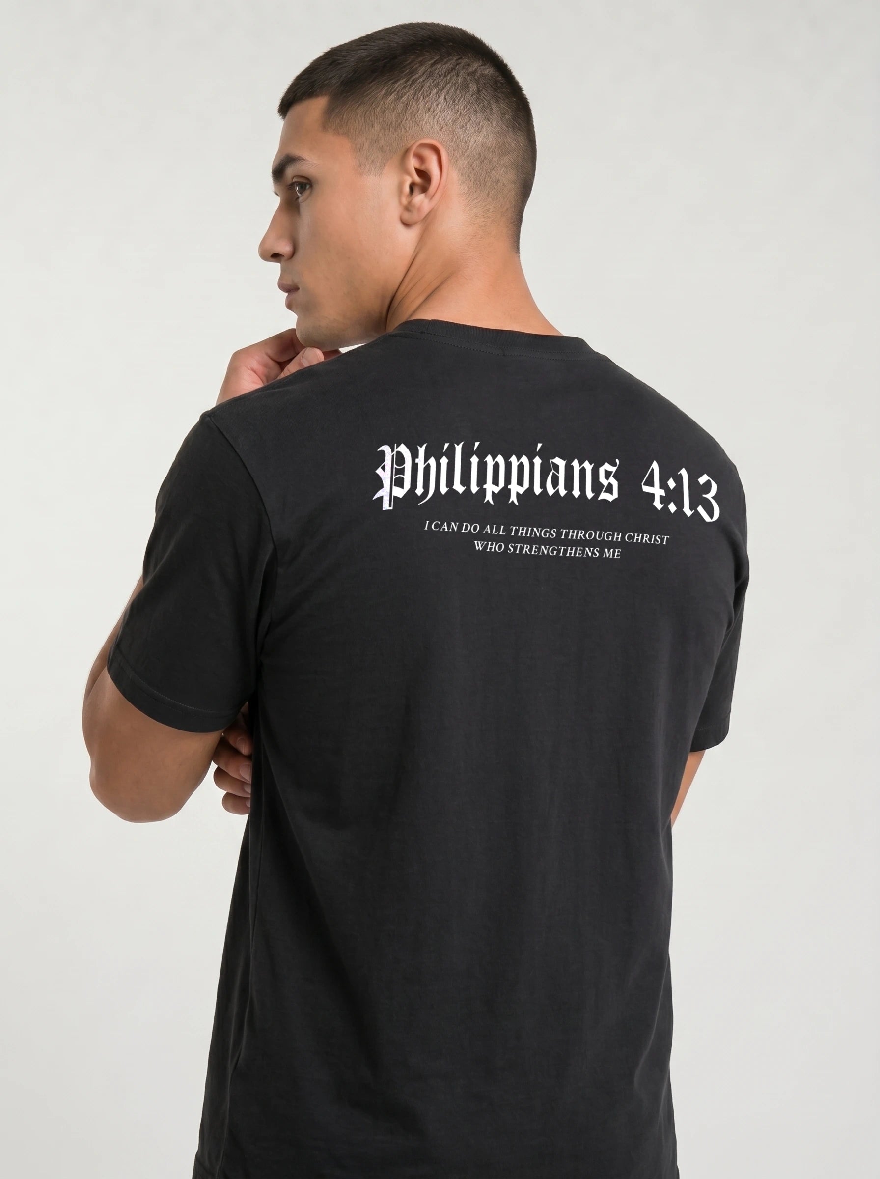 A man wearing a black t-shirt, viewed from the back. The shirt features the text 'Philippians 4:13' in a white Gothic font, with the subtitle 'I CAN DO ALL THINGS THROUGH CHRIST WHO STRENGTHENS ME' printed below in smaller white text.