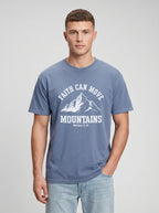 A man wearing a slate blue crewneck t-shirt with a white graphic print that reads 'FAITH CAN MOVE MOUNTAINS' and 'Matthew 17:20' below a mountain illustration, paired with light wash jeans against a light gray studio background.