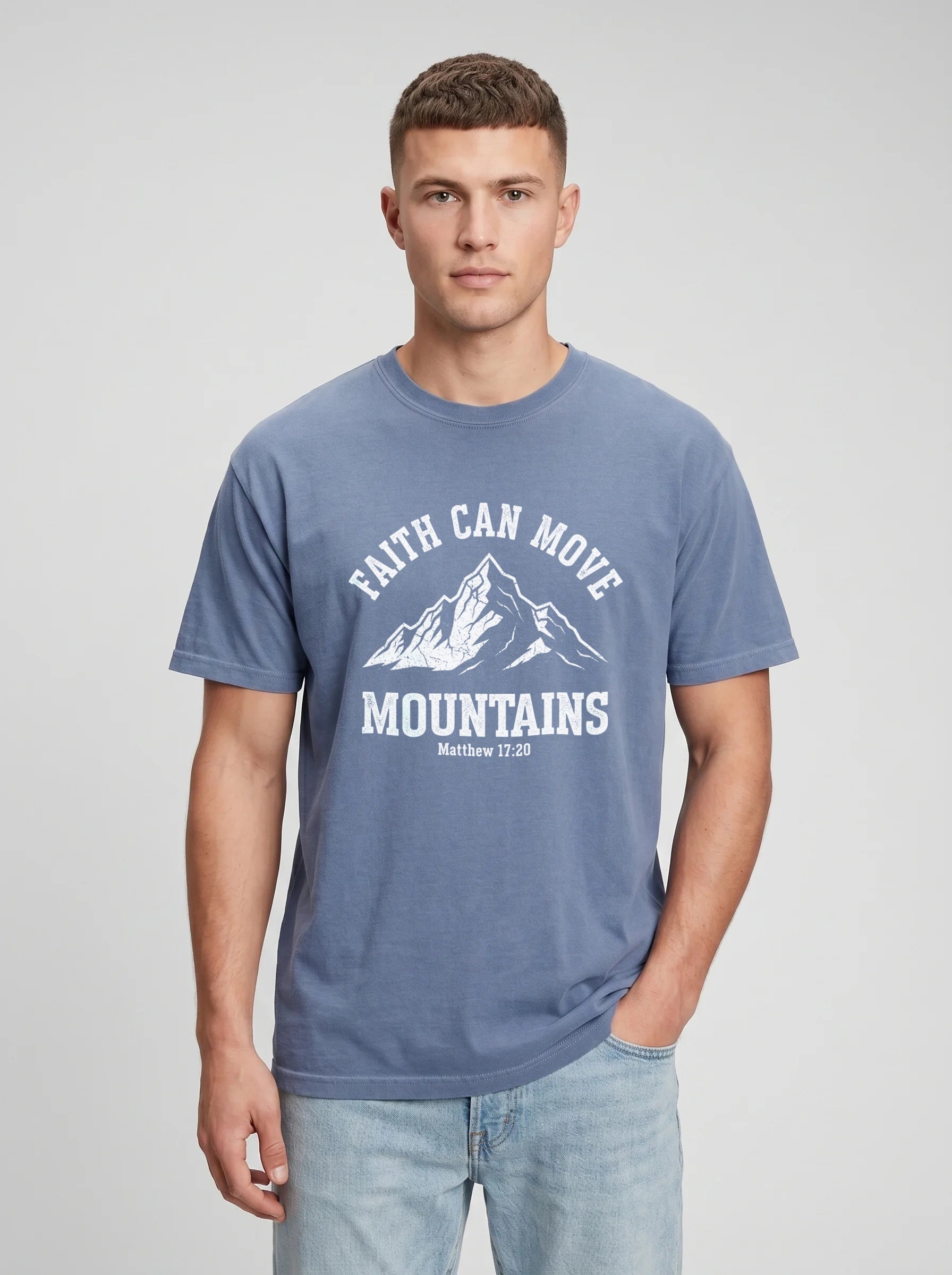 A man wearing a slate blue crewneck t-shirt with a white graphic print that reads 'FAITH CAN MOVE MOUNTAINS' and 'Matthew 17:20' below a mountain illustration, paired with light wash jeans against a light gray studio background.