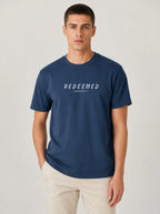 A man wearing a navy blue Comfort Colors t-shirt with "REDEEMED" in bold white capital letters and "EPHESIANS 1:7" underneath, paired with beige pants, against a plain light background.