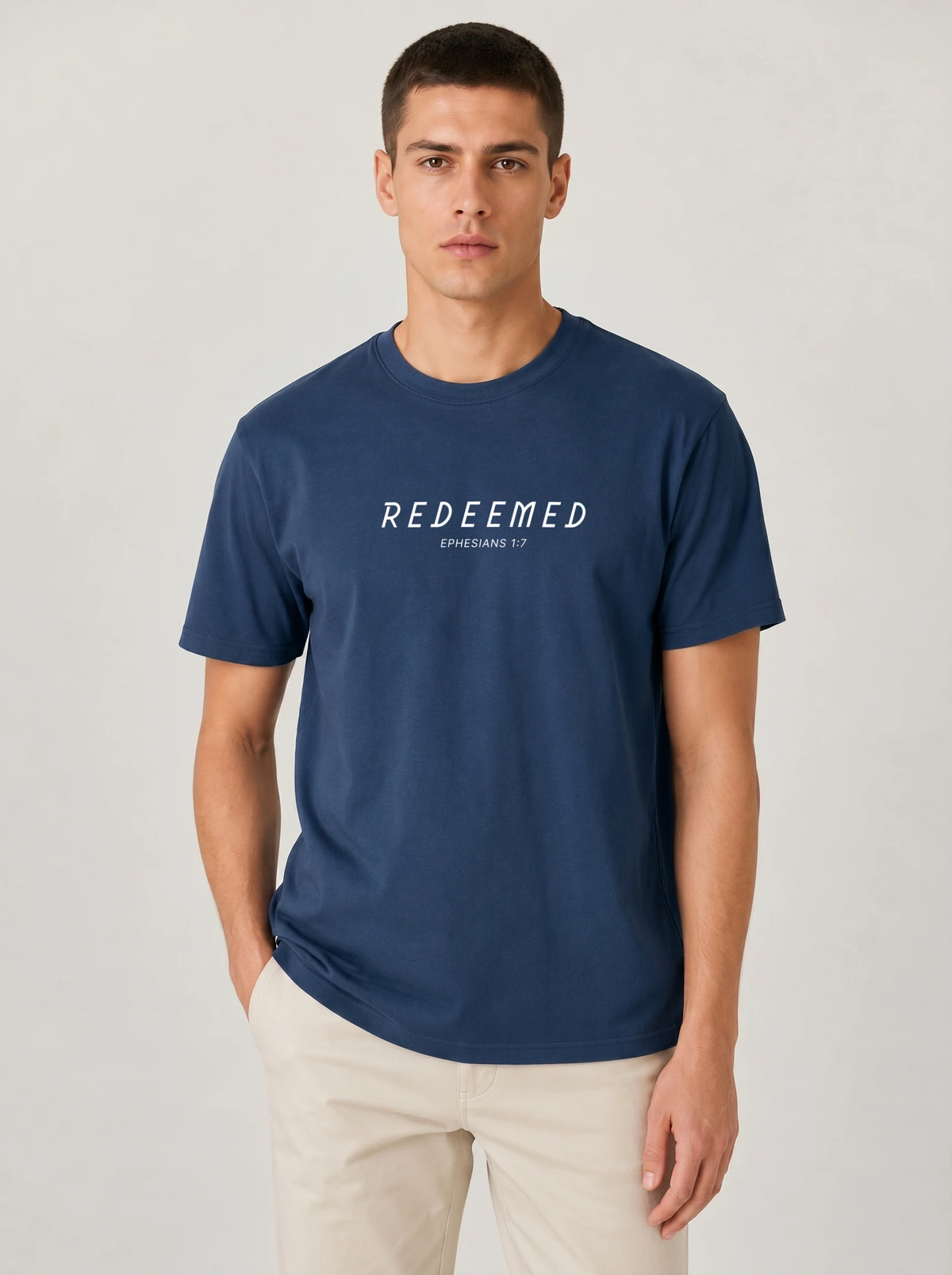 A man wearing a navy blue Comfort Colors t-shirt with "REDEEMED" in bold white capital letters and "EPHESIANS 1:7" underneath, paired with beige pants, against a plain light background.