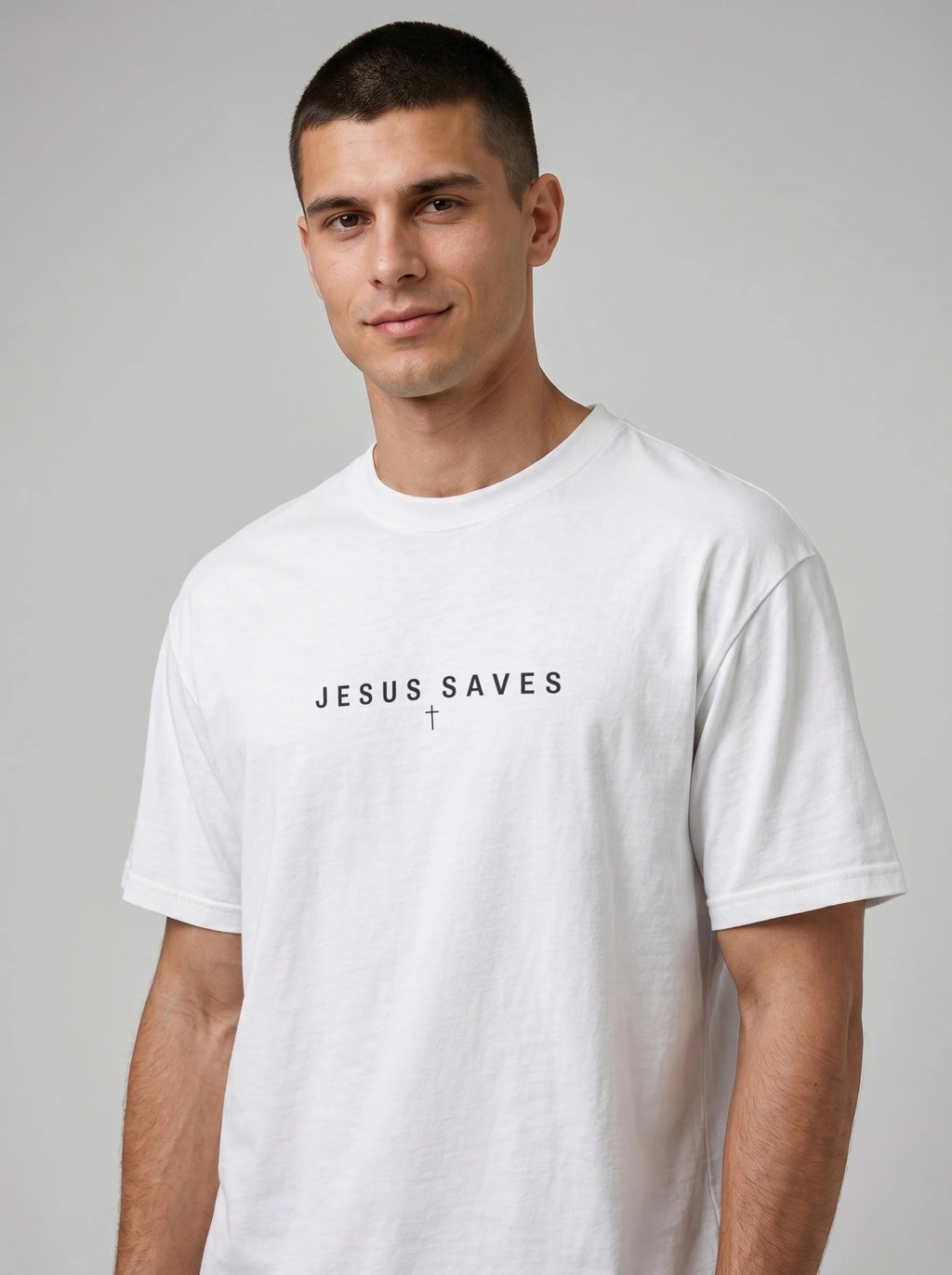 A man wearing a white crew-neck t-shirt with the text 'JESUS SAVES' and a small cross printed in black on the chest, photographed against a neutral gray background.