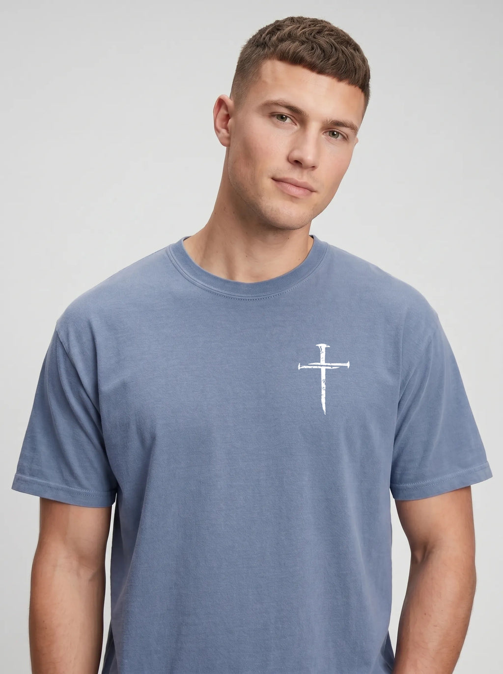 A man wearing a heathered slate blue t-shirt with a distressed nail cross on his left chest, photographed against a neutral light gray background.