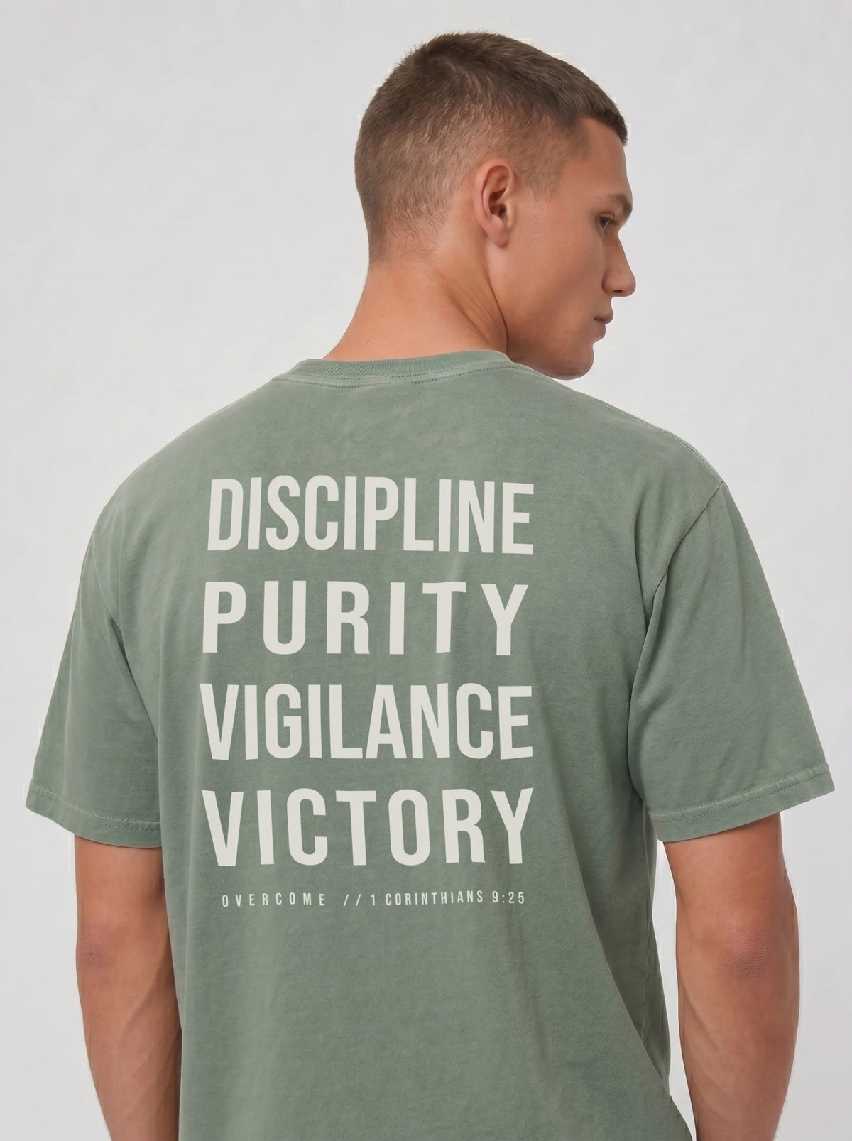 Person wearing a muted green T-shirt with large back text reading “Discipline Purity Vigilance Victory” and “Overcome // 1 Corinthians 9:25.”