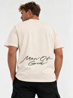 Person wearing a cream T-shirt with a large black script graphic across the lower back reading “Man Of God.”
