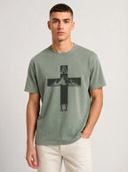 A man wearing a sage green crewneck t-shirt with a black cross graphic featuring a mountain and the letters A and Omega, paired with off-white pants, against a light gray studio background.