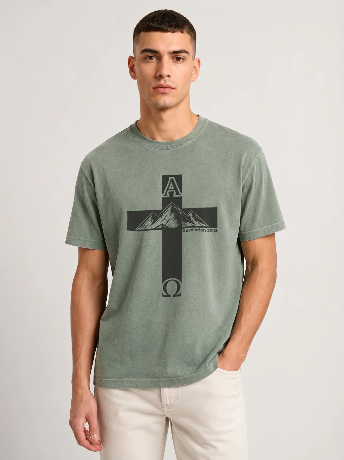A man wearing a sage green crewneck t-shirt with a black cross graphic featuring a mountain and the letters A and Omega, paired with off-white pants, against a light gray studio background.