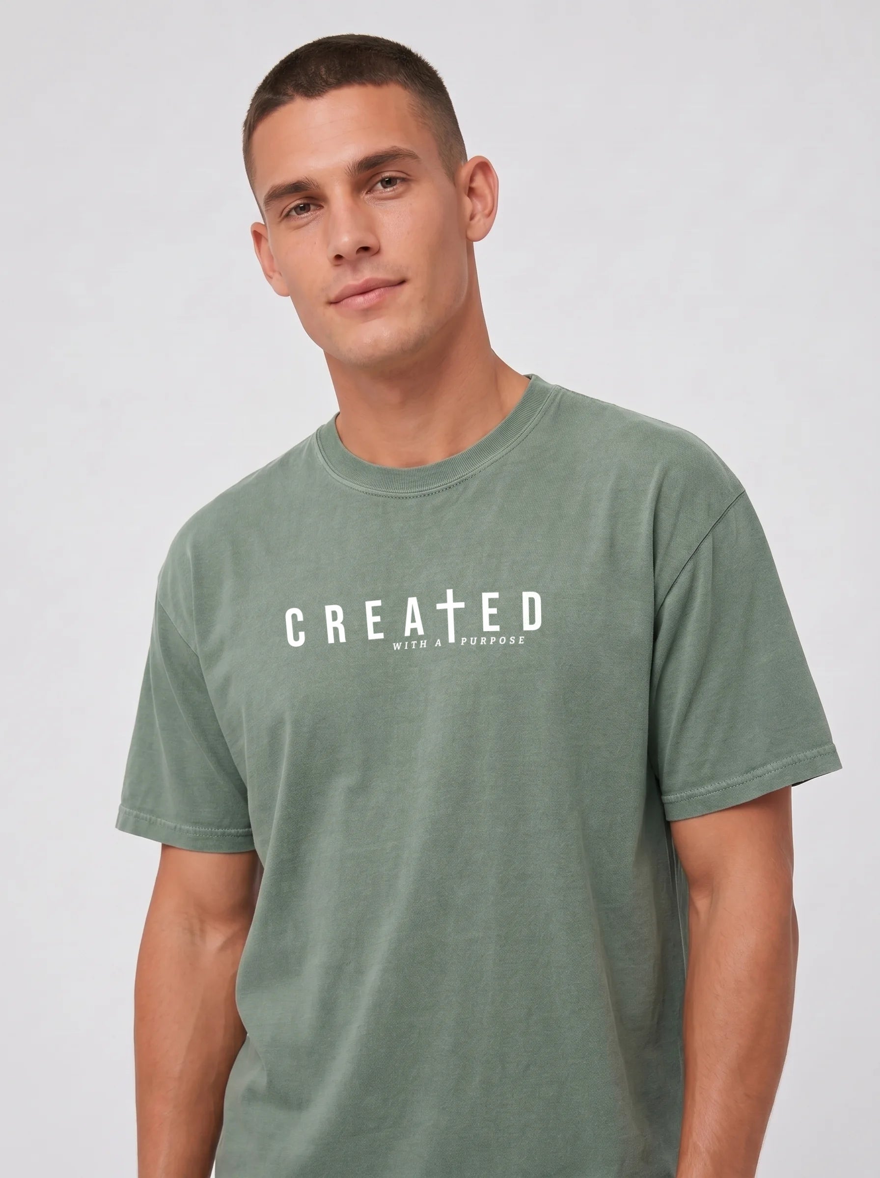 A man wearing a muted sage green crewneck t-shirt with 'CREATED with a purpose' printed in white across the chest, set against a plain light gray studio background.