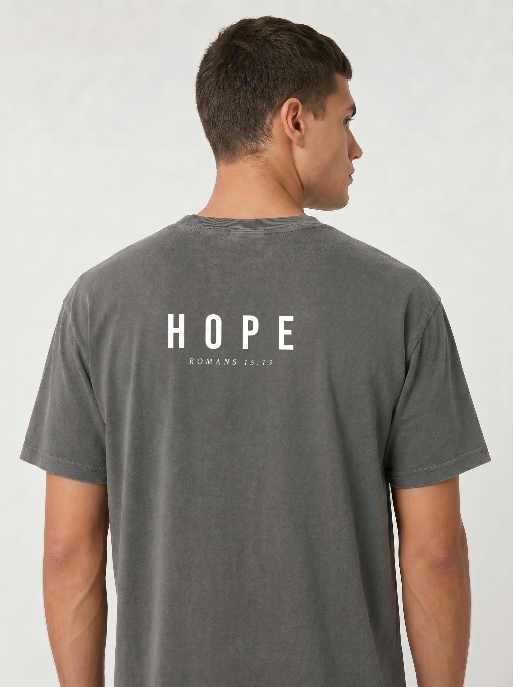 A man viewed from the back wearing a dark gray t-shirt with 'HOPE ROMANN 15:13' printed in white on the back, set against a light gray studio background.