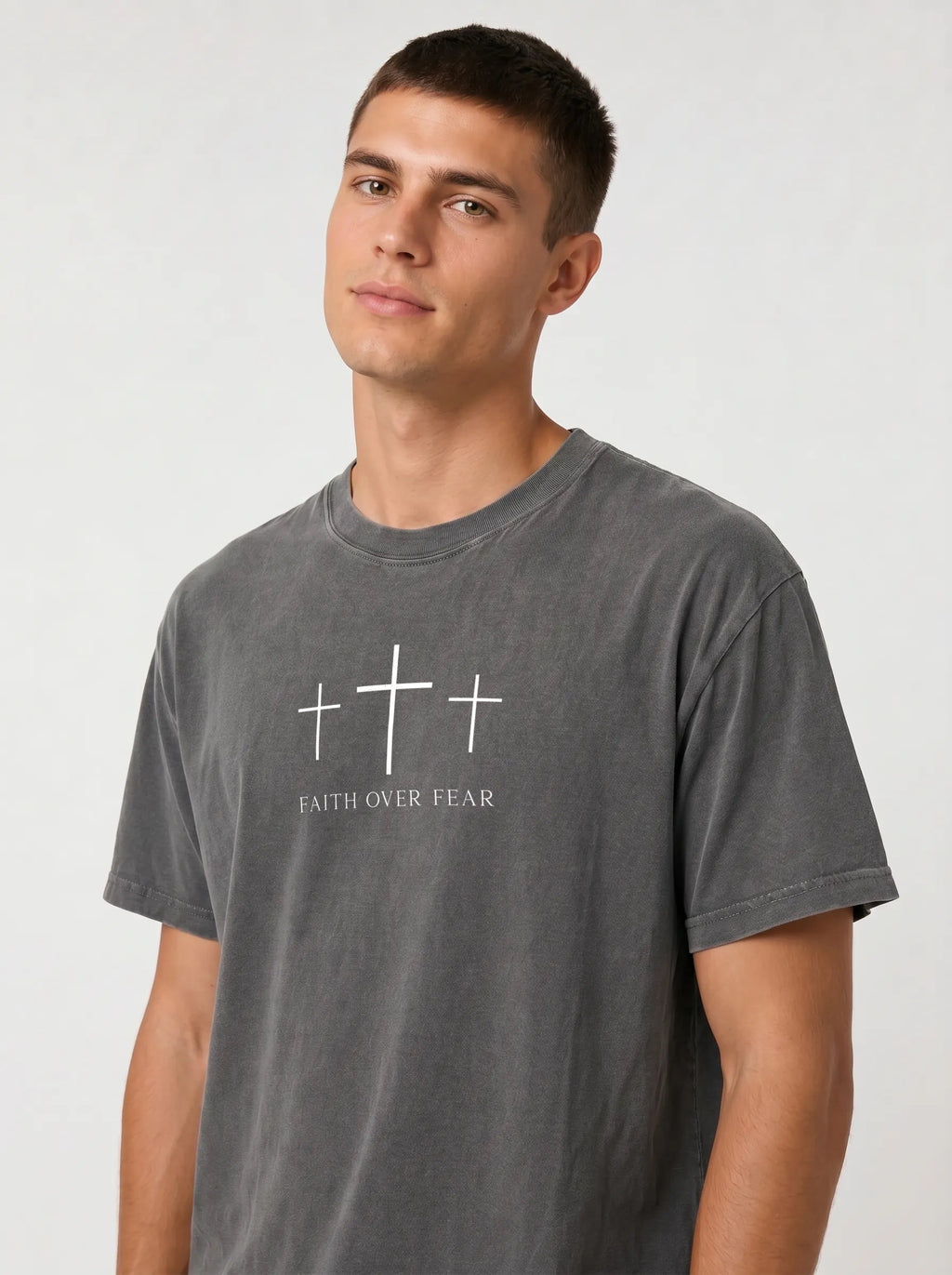 A man wearing a dark gray washed t-shirt with a white graphic of three crosses and the text 'FAITH OVER FEAR' printed on the chest, set against a plain light gray background.