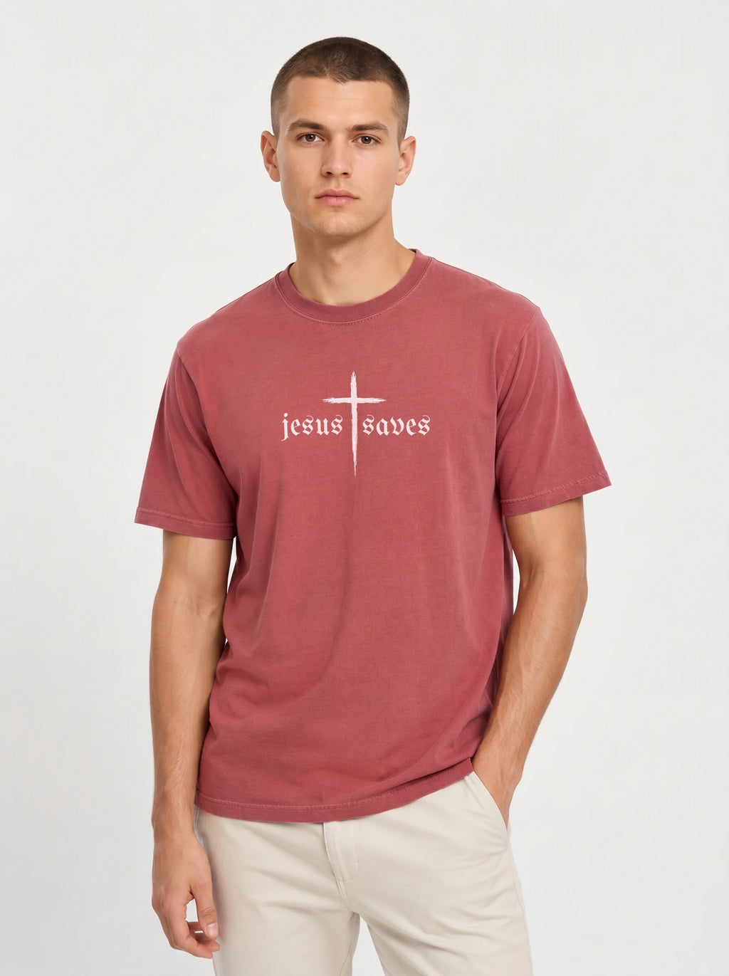 A man wearing a dusty rose short-sleeve t-shirt with a white gothic-style cross and 'jesus saves' text on the chest, paired with light beige pants, against a light gray studio background.